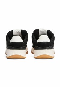 Zapatillas en blanco y negro con parte superior de ante, cuello acolchado y suela de goma beige. Presenta detalles de la marca en el talón.