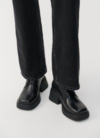 Botas de couro pretas com um salto grosso, design de atacadores e biqueira arredondada. Combinadas com jeans pretos de perna larga, mostrando linhas limpas e textura.