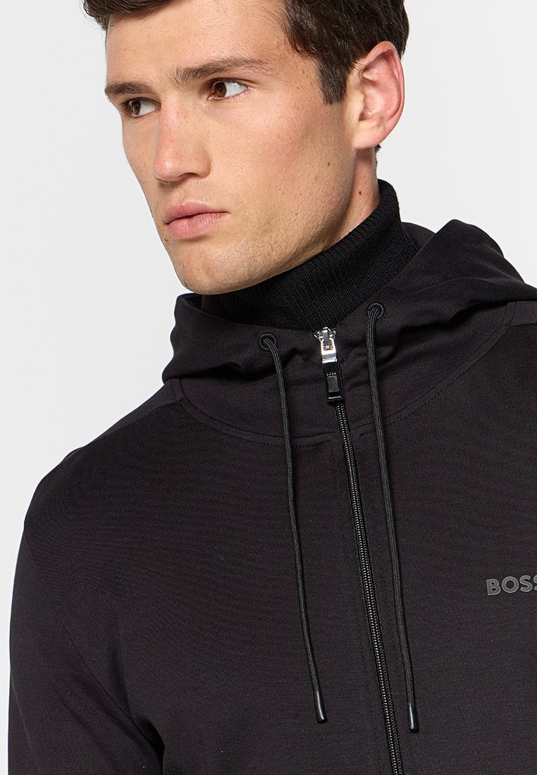 Jonge man in een zwart ritsvesting over een zwarte coltrui, kijkend naar de zijkant tegen een effen lichte achtergrond.