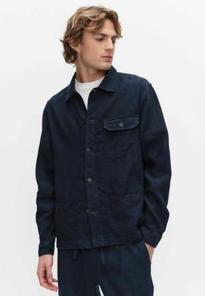 Nuori mies aaltoilevilla hiuksilla, pukeutuneena tummansiniseen napitettuun takkiin, jossa on etutaskut, katse alaspäin ja vasemmalle yksiväristä taustaa vasten.