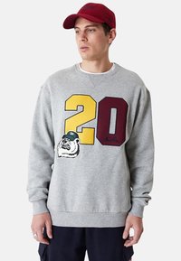 Sudadera gris con grandes números amarillos y marrones "20", un gráfico de un oso y puños y dobladillo acanalados. Ajuste casual con detalle bordado.