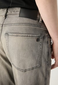 Jeans in denim grigio con una leggera texture sbiadita, dotati di una patch in pelle nera con il logo BOSS in vita e un dettaglio metallico sulla tasca posteriore.
