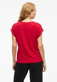 Maglietta rossa a maniche corte, in tessuto morbido, vestibilità comoda, scollatura rotonda, maniche arrotolate, texture liscia, indossata con pantaloni neri.