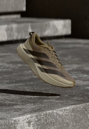 Chaussure de course marron clair avec des rayures foncées et une semelle épaisse flottant au-dessus de marches en pierre texturée.