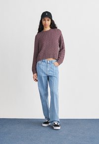 Νέα γυναίκα που φοράει μωβ πλεκτό πουλόβερ με κοτσίδες, ριγέ ανοιχτό μπλε τζιν, μαύρο καπέλο και μαύρα αθλητικά παπούτσια, στέκεται σε μπλε χαλί.