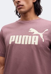 Κοντινό πλάνο ενός ατόμου που φοράει μωβ T-shirt με μεγάλο λευκό λογότυπο Puma και γραφικό πετάγματος πούμα στο στήθος.