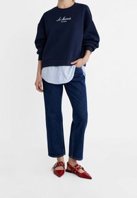 Felpa navy con testo "Le Marais Paris", sovrapposta a una camicia a righe blu chiaro, abbinata a jeans denim scuri e ballerine rosse a punta.