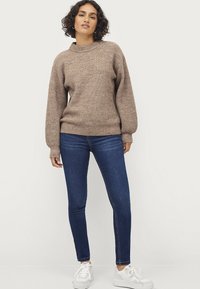 Maglione a maglia marrone con scollatura alta, texture a coste e maniche lunghe, abbinato a jeans skinny blu e sneakers bianche.