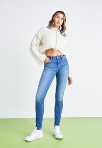 Femme portant un pull court blanc, un jean skinny bleu et des baskets blanches, debout sur un sol vert devant un fond blanc.