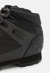Scarponi da trekking neri con tomaia sintetica texturizzata, collo imbottito e suola in gomma con dettagli a pattern. Logo Timberland visibile.