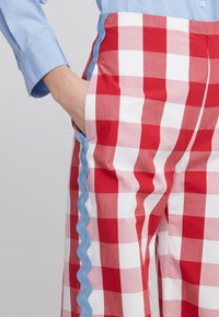 Punat punut punja dengan desain kotak-kotak merah dan putih, dilengkapi dengan trim scalloped biru yang dijahit di samping, terbuat dari kain ringan, menampilkan potongan longgar dan saku.