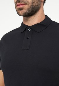 Homme portant un polo noir à deux boutons, montrant la partie inférieure du visage et le cou avec une barbe courte et une moustache.