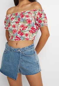 Donna che indossa un top a spalle scoperte floreale con fiori rossi, verdi e blu abbinato a una mini gonna in denim asimmetrica.