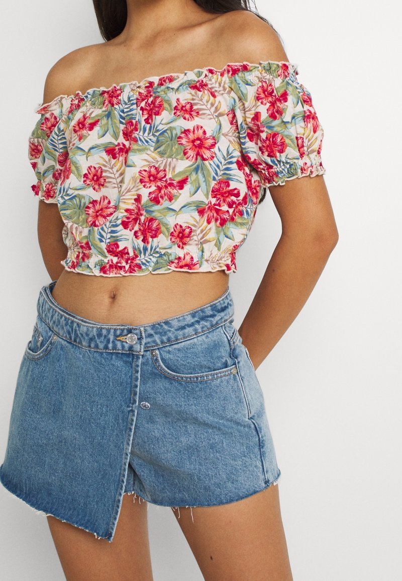 Donna che indossa un top a spalle scoperte floreale con fiori rossi, verdi e blu abbinato a una mini gonna in denim asimmetrica.