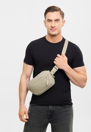 Bărbat în tricou negru și blugi închiși la culoare ținând o curea bej de geantă crossbody pe fundal alb.