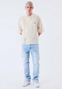Korte mouwen, lichtgele knoopsluiting met een gestructureerd patroon, gecombineerd met lichtblauwe versleten jeans en witte sneakers.