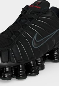 Čierna bežecká obuv Nike Shox s lesklou podrážkou pohlcujúcou nárazy, našitým logom Swoosh a červeným textom "Nike Shox" na jazyku.