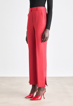 Pantalons rouges ajustés avec une taille haute, des fentes sur les côtés et des passants de ceinture, associés à des chaussures à talons hauts rouges brillantes. Le tissu semble lisse.