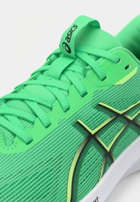 Zapatilla deportiva verde con parte superior de malla texturizada, cordones planos, detalles en amarillo y suela blanca. Presenta un destacado diseño de logo negro y verde.