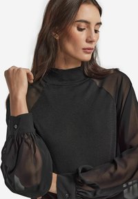 Top nero con colletto alto, caratterizzato da maniche a palloncino trasparenti. Il corpo è texturizzato, mentre le maniche sono lisce e fluenti, presentando un design a strati.