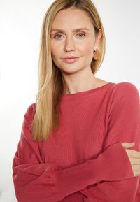 Pulover roz, cu mâneci lungi, cu textură în vrac și manșete ample. Modelul poartă cercei rotunzi și transparenți. Fundalul deschis pune accent pe articolul de îmbrăcăminte.