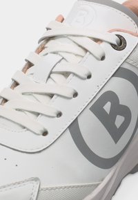 Gros plan sur une basket blanche avec des lacets blancs, un logo "B" gris sur le côté, et un œillet métallique près du col rembourré avec une doublure intérieure pêche.