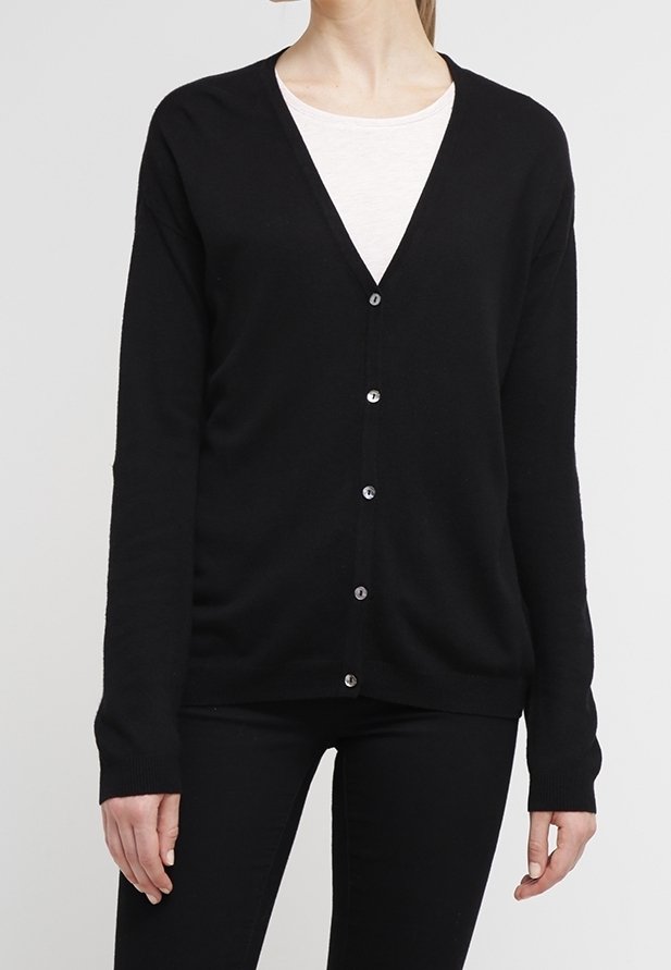 Femme portant un cardigan noir boutonné par-dessus un haut blanc et un pantalon noir, debout devant un fond gris clair.