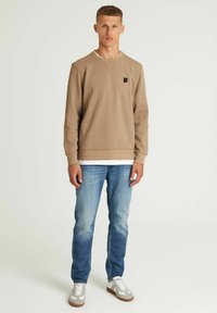 Sudadera marrón claro con puños acanalados, con un pequeño logo negro en el pecho. Combinada con unos vaqueros azules desgastados y zapatillas grises.