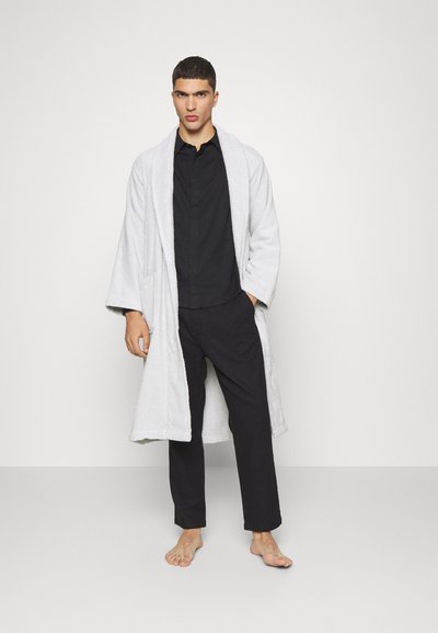 Robe de toalha cinza claro com mangas longas e bolsos frontais, usado sobre uma camisa preta com botões e calças pretas, exibido por um modelo.