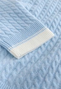 Manche de pull bleu clair à maille torsadée avec un poignet en côte blanc orné d'une fine rayure bleue, présenté sur un fond en tissu assorti.