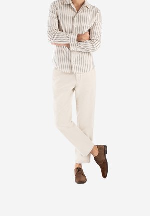 Chemise rayée à manches longues en beige et marine, associée à un pantalon beige et des chaussures en daim marron, arborant un design épuré et une coupe ajustée.