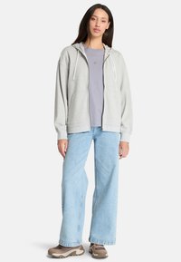 Haut à zip gris avec une capuche à cordon, associé à un t-shirt violet clair et un jean ample bleu clair. Chaussures neutres notables.