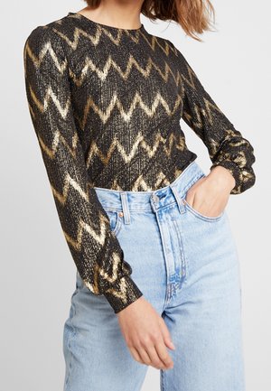 Femme portant un haut à manches longues noir avec un motif zigzag doré et un jean taille haute bleu clair, main dans la poche sur fond uni.