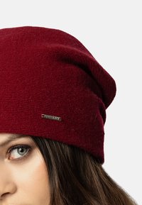 Burgundská pletená čepice s volnějším designem, s malým kovovým logem a měkkou texturou. Detailní záběr čepice blízko oka.