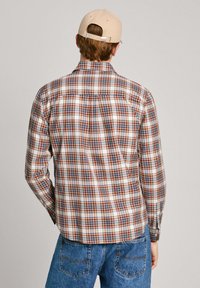Camisa de manga larga a cuadros en tonos beige, marrón y azul; cuenta con una costura en la sisa y puños de botón. Llevada con pantalones de mezclilla en azul claro.