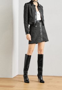 Chaqueta de tweed negra con detalles de cuero, combinada con una falda de tweed a juego con detalles de botones. Botas de cocodrilo negras de charol hasta la rodilla.