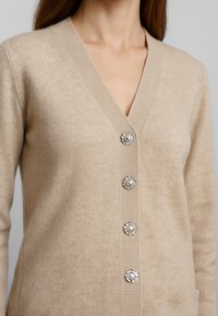 Cardigan beige realizzato in morbido tessuto a maglia, con design a V, cinque bottoni decorativi a gemma e dettagli a costine lungo i bordi.