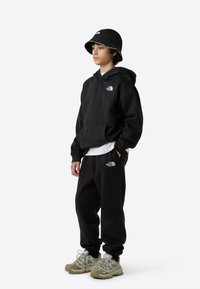 Hanorac și pantaloni de trening negri cu logo alb, asortați cu o pălărie tip bucket neagră. Pantofi sport gri și bej, cu design din plasă și talpă texturată.