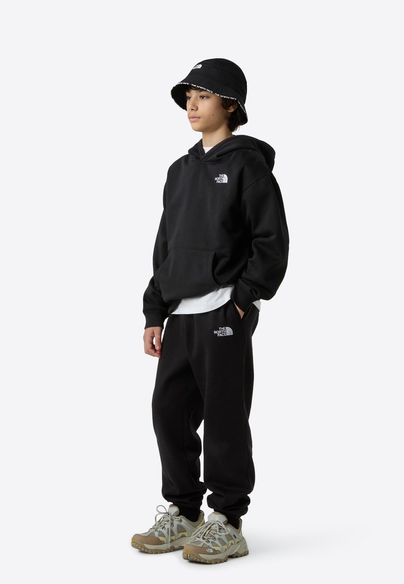 Hanorac și pantaloni de trening negri cu logo alb, asortați cu o pălărie tip bucket neagră. Pantofi sport gri și bej, cu design din plasă și talpă texturată.