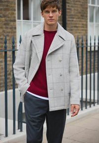 Harmaa ruudullinen merimiestakki leveällä kauluksella, kaksirivinen etuosa, yhdistetty burgundinväriseen villapaitaan ja tummanharmaisiin housuihin. Sileä rakenne, rento istuvuus.