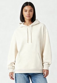 Felpa con cappuccio color crema in tessuto morbido, con cappuccio regolabile a coulisse, tasca frontale e polsini a coste. Indossata con jeans blu.