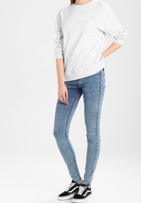 Šedá mikina s kulatým výstřihem a texturovanou bílou grafikou; spárována se světle modrými slim fit džíny a černými nízkými teniskami.