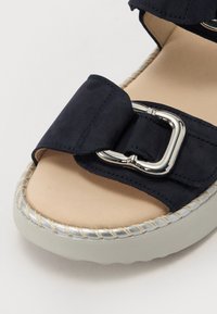 Navy blue suede sandal featuring a large buckle, beige insole, and a rounded grey sole with a woven rope accent along the edge.