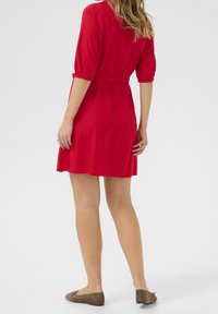 Czerwona sukienka na krótki rękaw z dopasowaną talią, zwiewnym dołem i paskiem. Płaskie brązowe buty dopełniają wygląd. Gładki materiał z minimalnymi detalami.