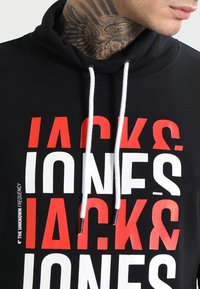 Felpa nera con cappuccio, con grande scritta "JACK & JONES" in rosso e bianco. I cordini sono bianchi con punte in metallo.