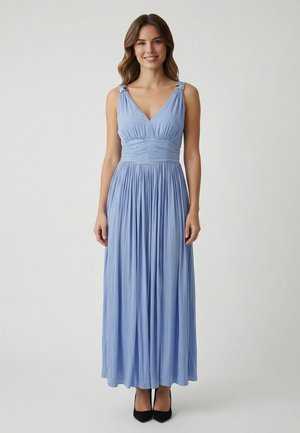 Femme debout portant une robe maxi sans manches bleu pâle avec des plis et une taille froncée ajustée, chaussée de talons noirs à bout pointu.