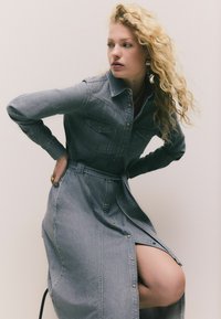 Denimkleid in Hellblau mit durchgehender Knopfleiste, langen Ärmeln und zwei Brusttaschen. Mit tailliertem Gürtel und seitlichem Schlitzdetail.