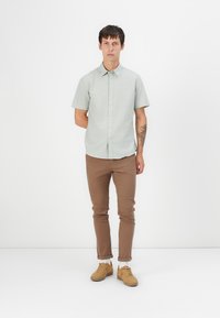 Camisa verde claro de manga corta con cuello abotonado, combinada con pantalones marrones ajustados y zapatillas color beige. Diseño simple y casual.