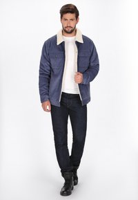 Tengerész kék kordbársony dzseki krémszínű gyapjú gallérral, két mellzsebbel és laza szabással, sötét farmernadrággal és fekete csizmával párosítva.