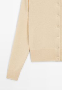 Beige gestrickte Strickjacke mit durchgehendem Knopfverschluss, gerippten Bündchen und Saum. Der Stoff hat eine weiche Textur mit einem dezenten Glanz und glatter Oberfläche.